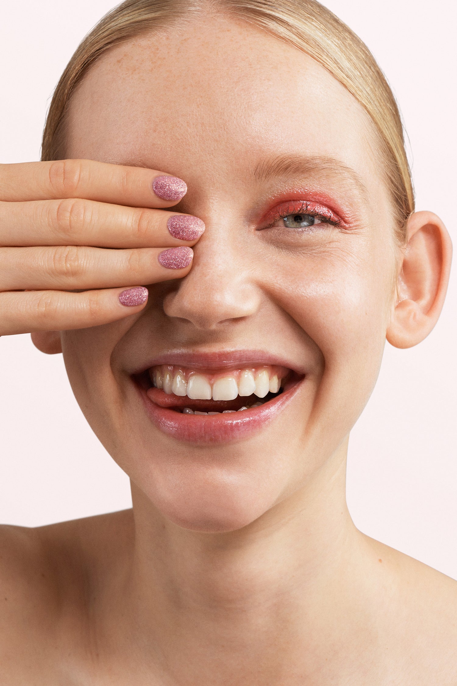 A smiling woman with pink eyeshadow covers an eye with her hand, which has pink glitter nail polish.