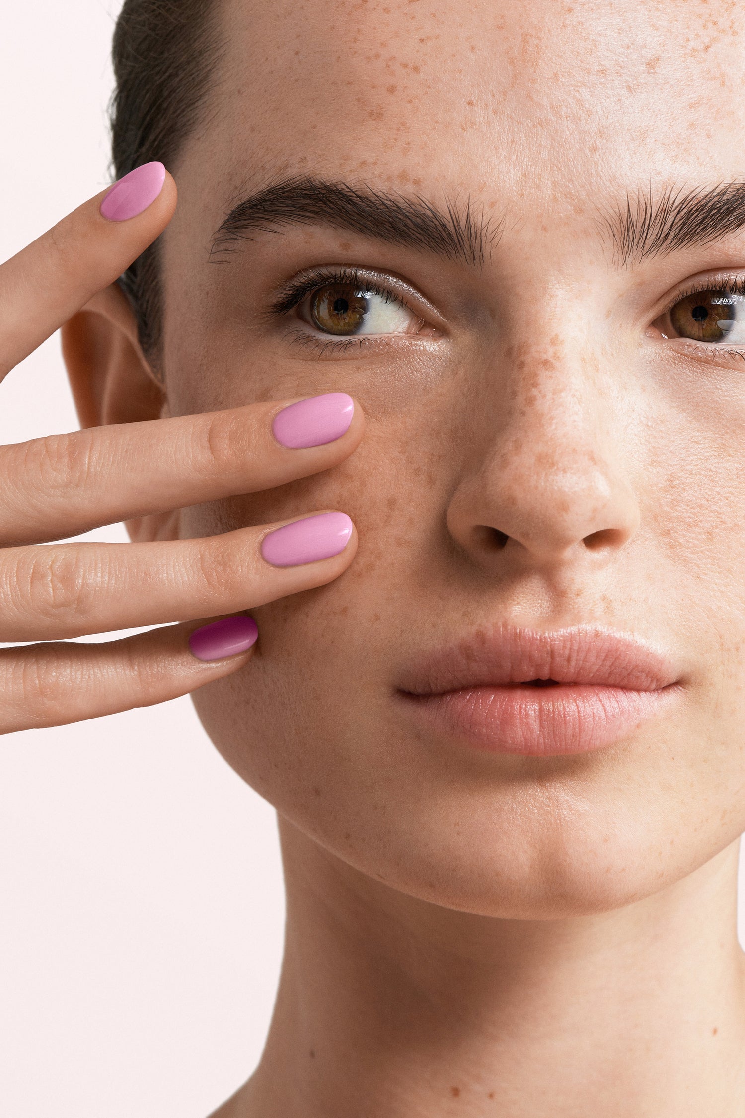 A close-up of a face with freckles and pink painted nails held against the cheek.