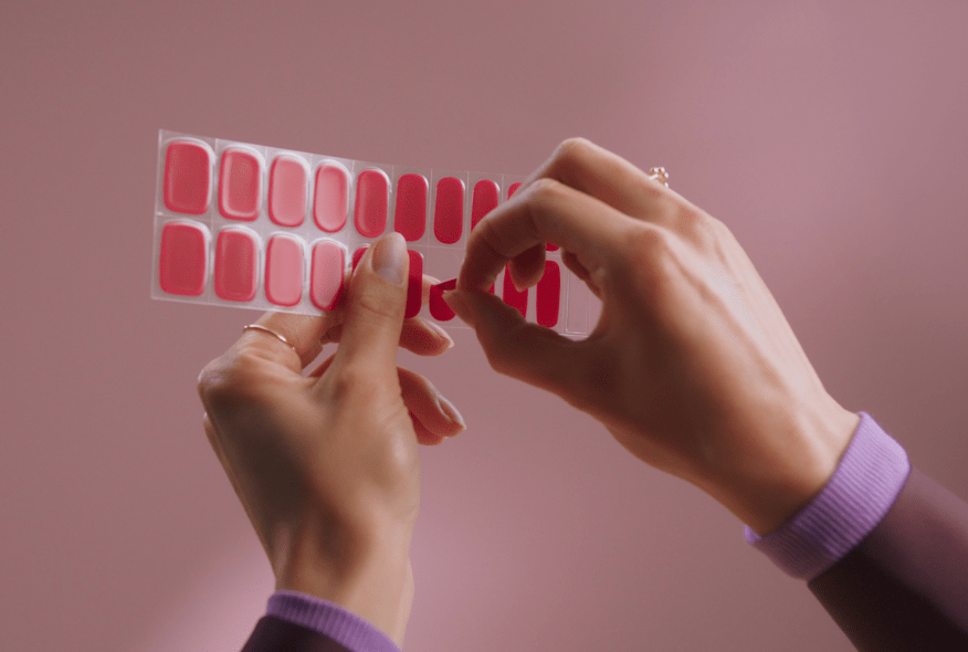 Two hands pulling pink nail stickers off a sheet against a pink background.
