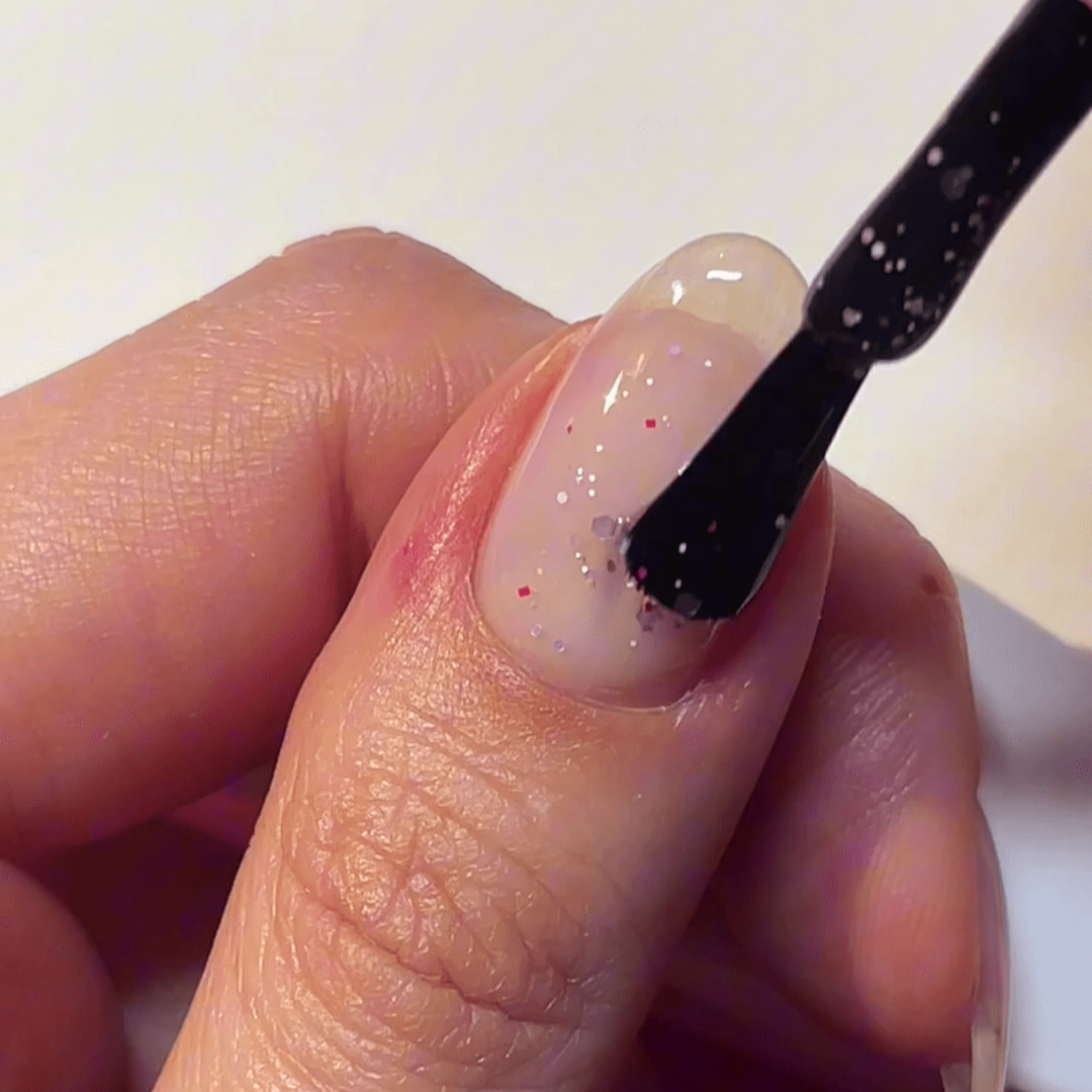 A hand applying nail polish with glitter to a nail.