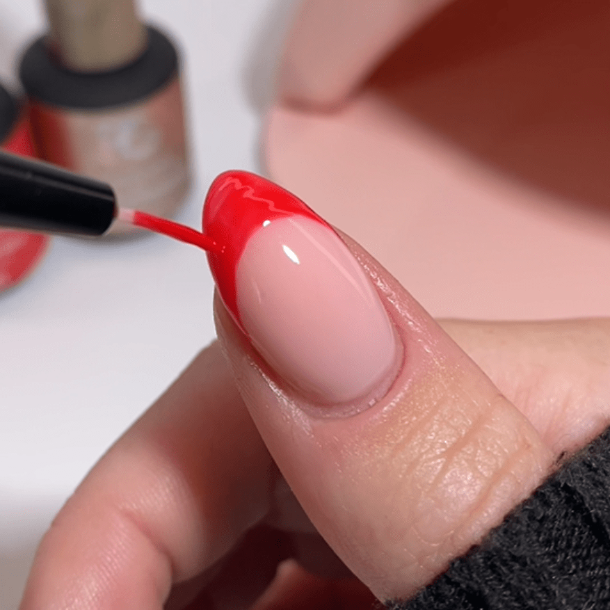 Close-up of a thumbnail being painted with bright red nail polish, while other nail polish bottles are visible in the background.