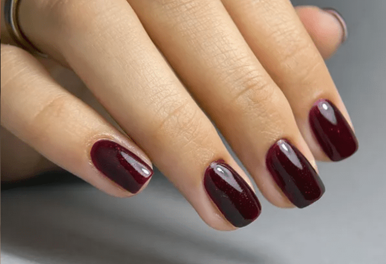 Hand holding short, dark red painted nails on a gray background.