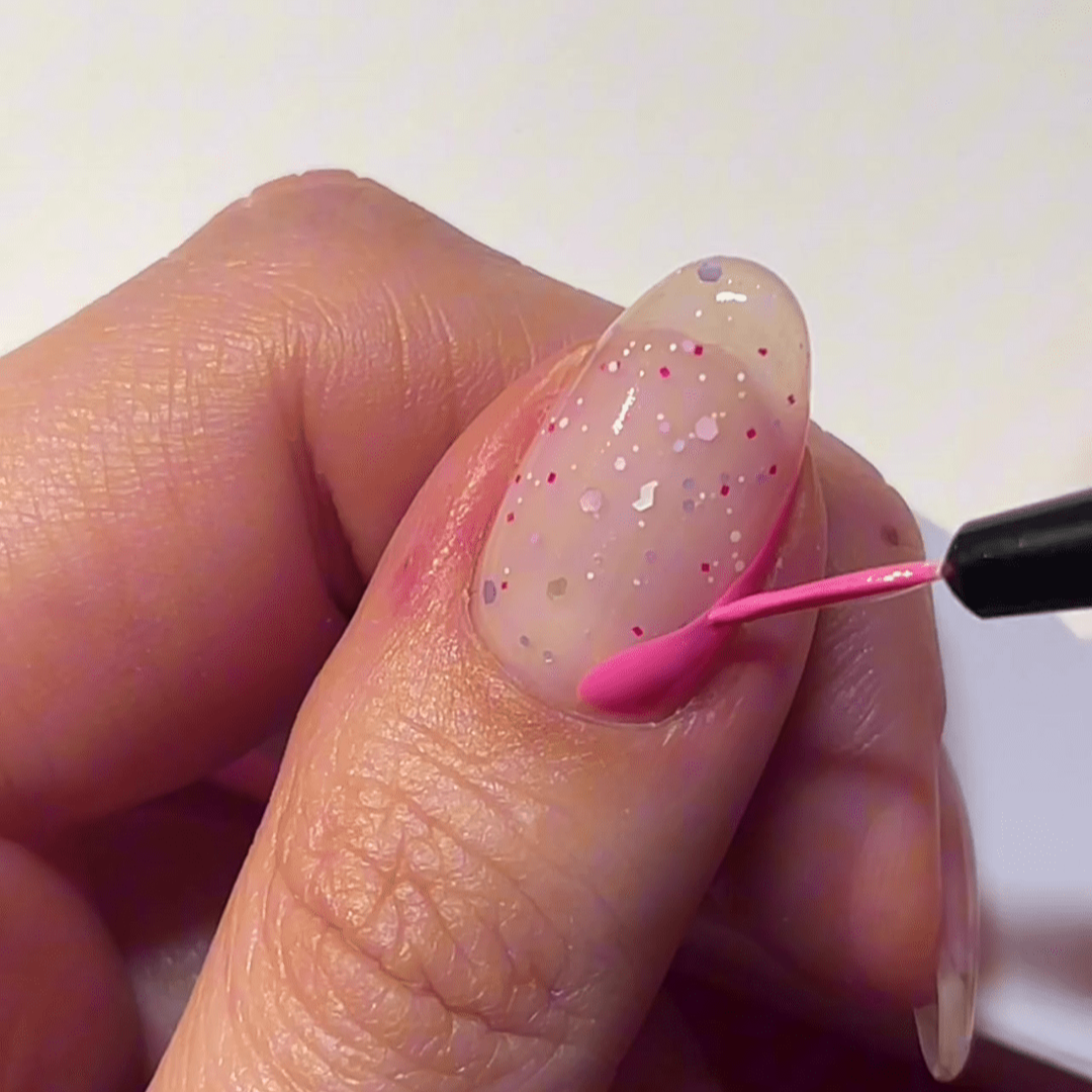 A hand applying a nail with pink and shimmering nail polish to a nail decorated with colorful dots.
