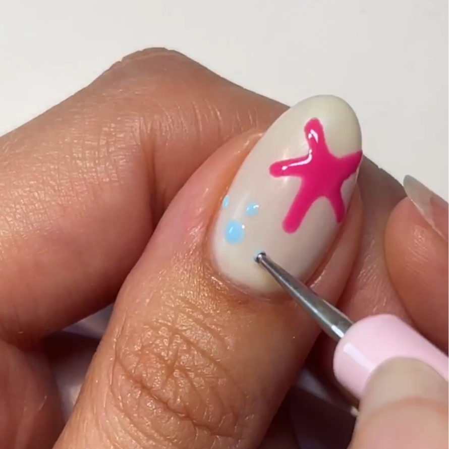 White nail with pink star shape and blue dots, hand with nail art tool.