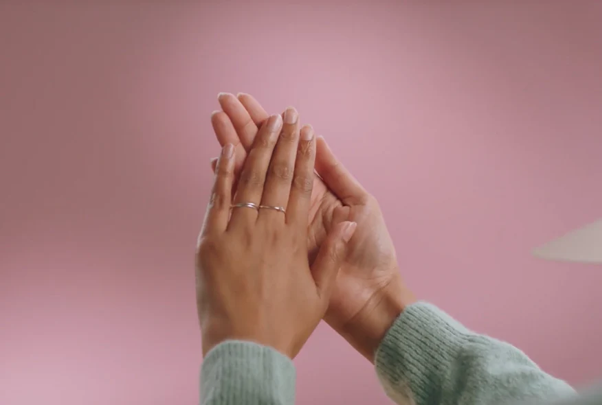 Two hands clap against a pink background; one wears silver rings, the other a green sweater.