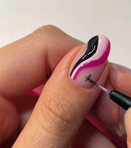 A close-up of a thumb with wavy pink, black and white nail art, on which a small black cross is applied with a brush.