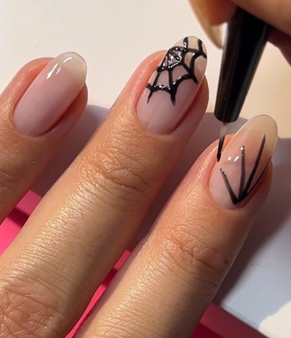 A hand with nails painted in a neutral color, where two nails are decorated with a black spider web pattern.
