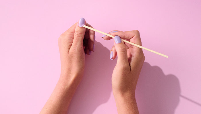 Two female hands with lilac painted nails take care of cuticles with a wooden stick against a pink background.