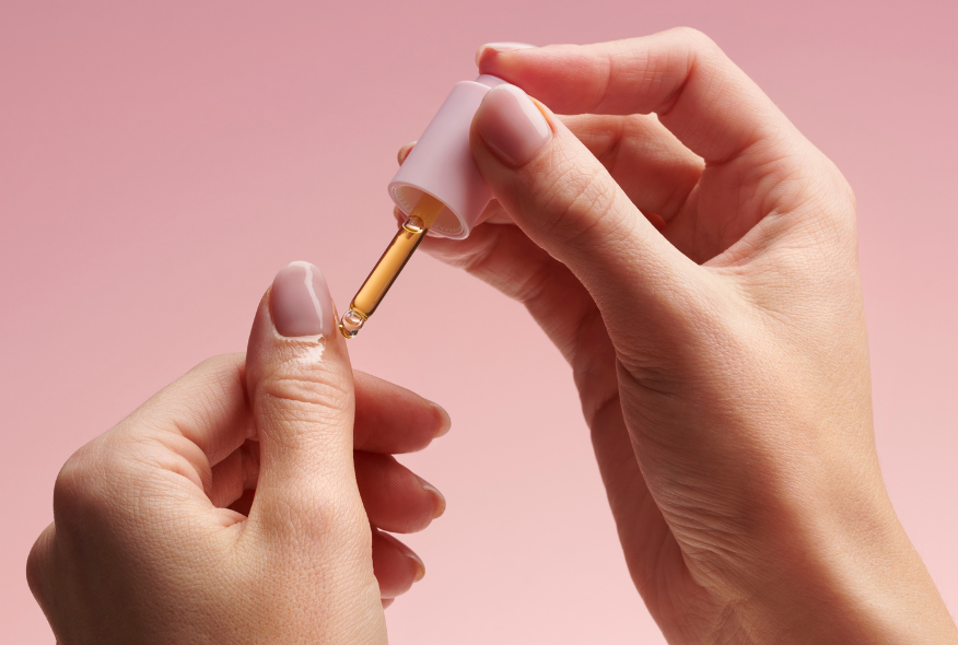 Hands holding a dropper with oil over a nail against a pink background.