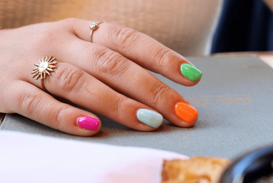 Hand with colorful nails and two graceful rings on a gray surface.