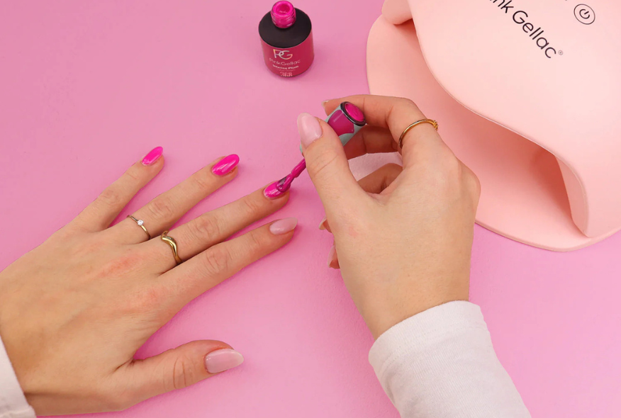 Hand painting nails with pink gel nail polish next to a UV lamp, on a pink surface.