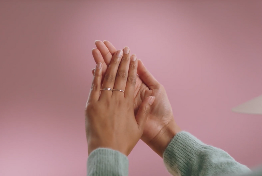 Two hands with fine rings press against each other with a pink background.