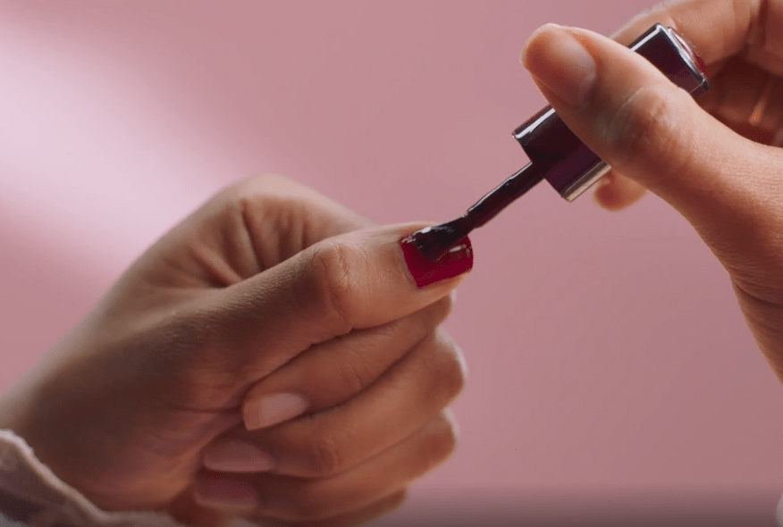 A person paints a nail with dark red nail polish against a pink background.