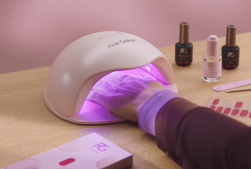 A hand is treated under a pink LED UV nail lamp from the Pink Gellac brand, surrounded by nail polish bottles and nail accessories on a wooden table.