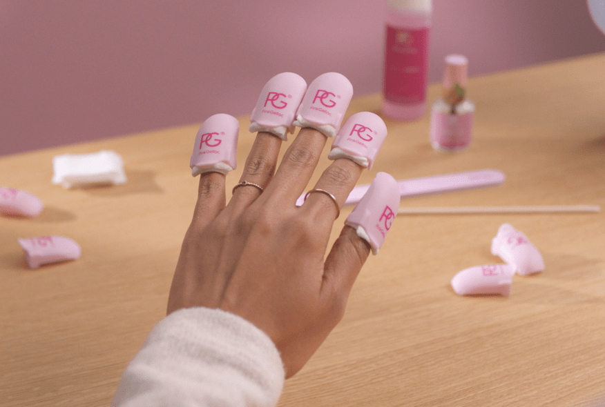 A hand with pink nail clips on each finger, surrounded by nail care products on a wooden table.