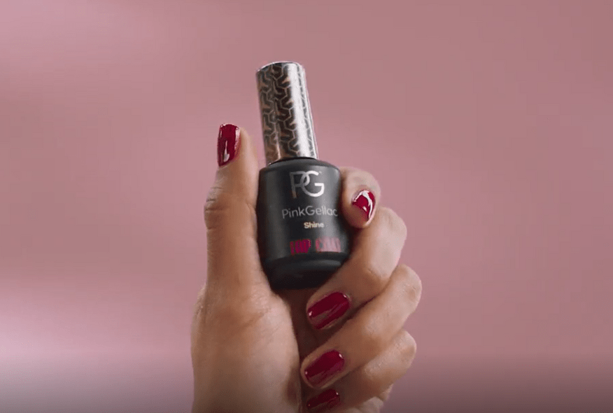A hand with wine-red nails holds a bottle of Pink Gellac nail polish, against a pink background.