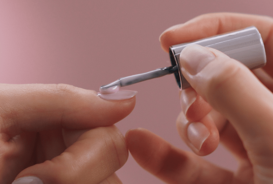 A person applies pink nail polish to a fingernail with a brush against a pink background.