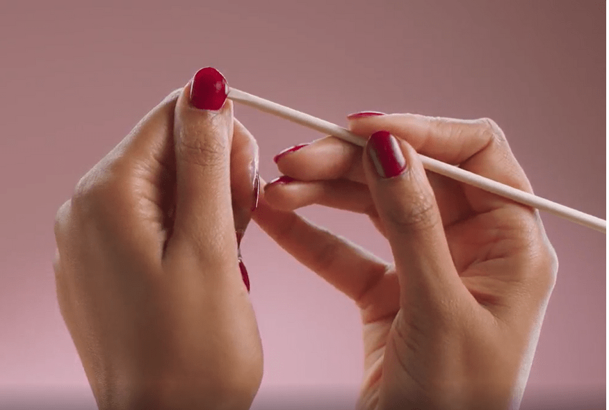 Hands with red nail polish holding a wooden stick for nail care against a pink background.