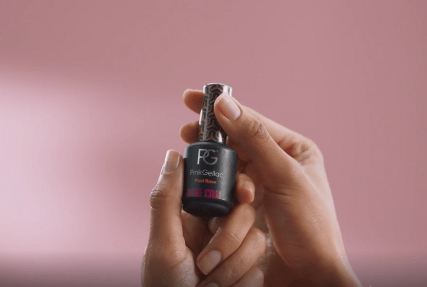 Two hands hold a bottle of Pink Gellac Peel Base nail polish against a pink background.