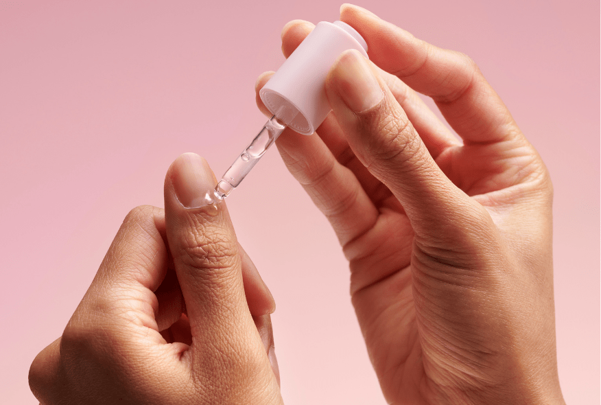 Hands using a dropper with liquid against a pink background.