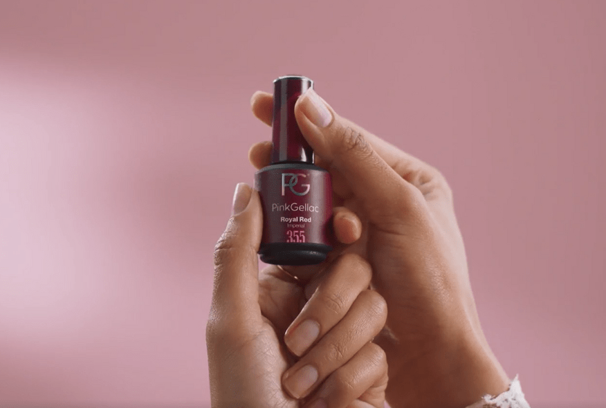 Two hands hold a bottle of dark red nail polish against a pink background.