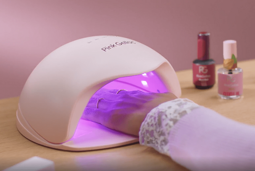 A hand under a UV lamp for drying nail polish, with two bottles of nail polish in the background.