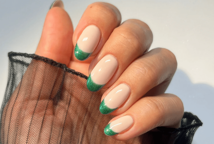 Hand with beige nails and green glitter tips, held against a black, transparent fabric.