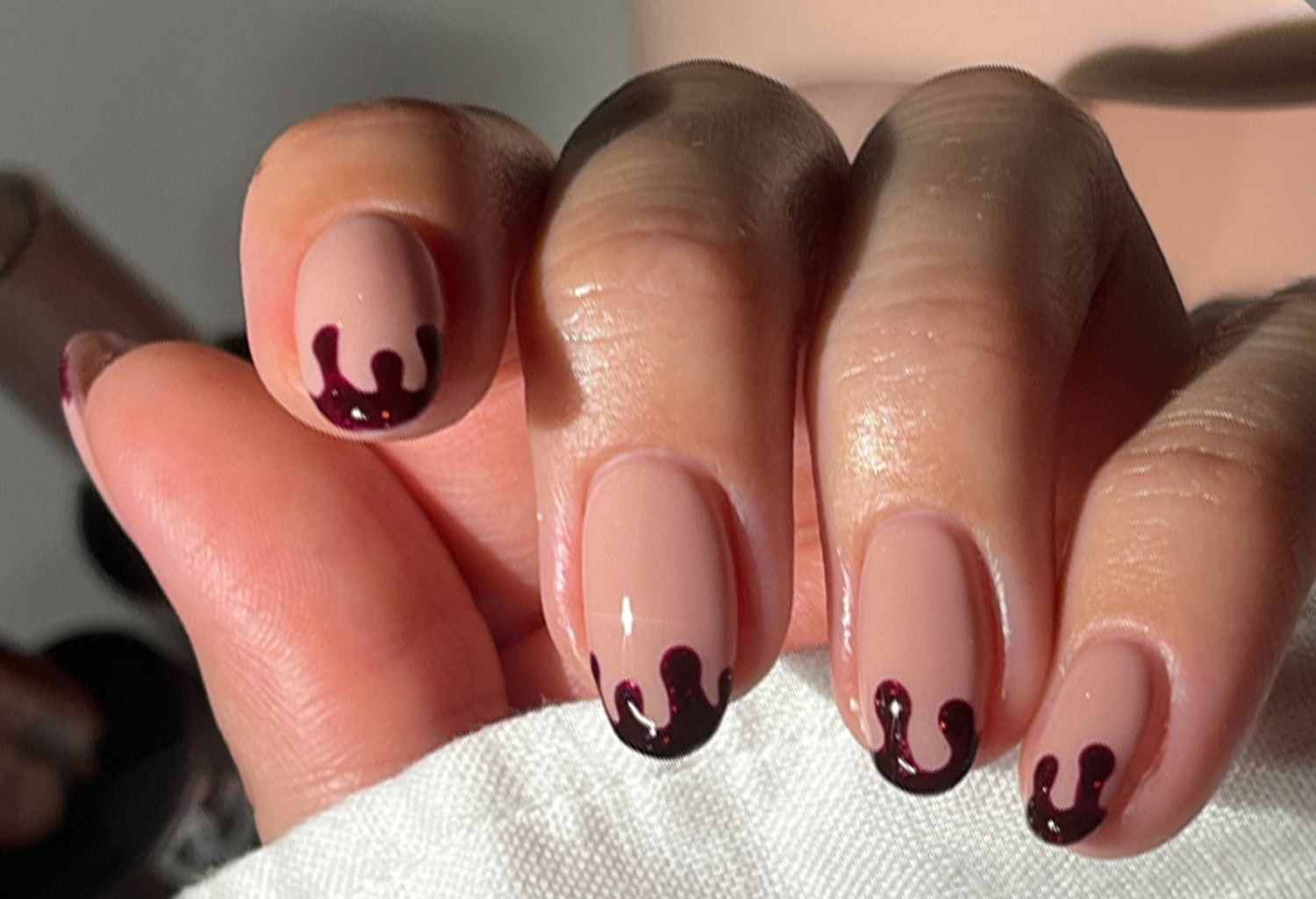 A hand with nails with a light pink base and dark red wavy tips, against a neutral background.