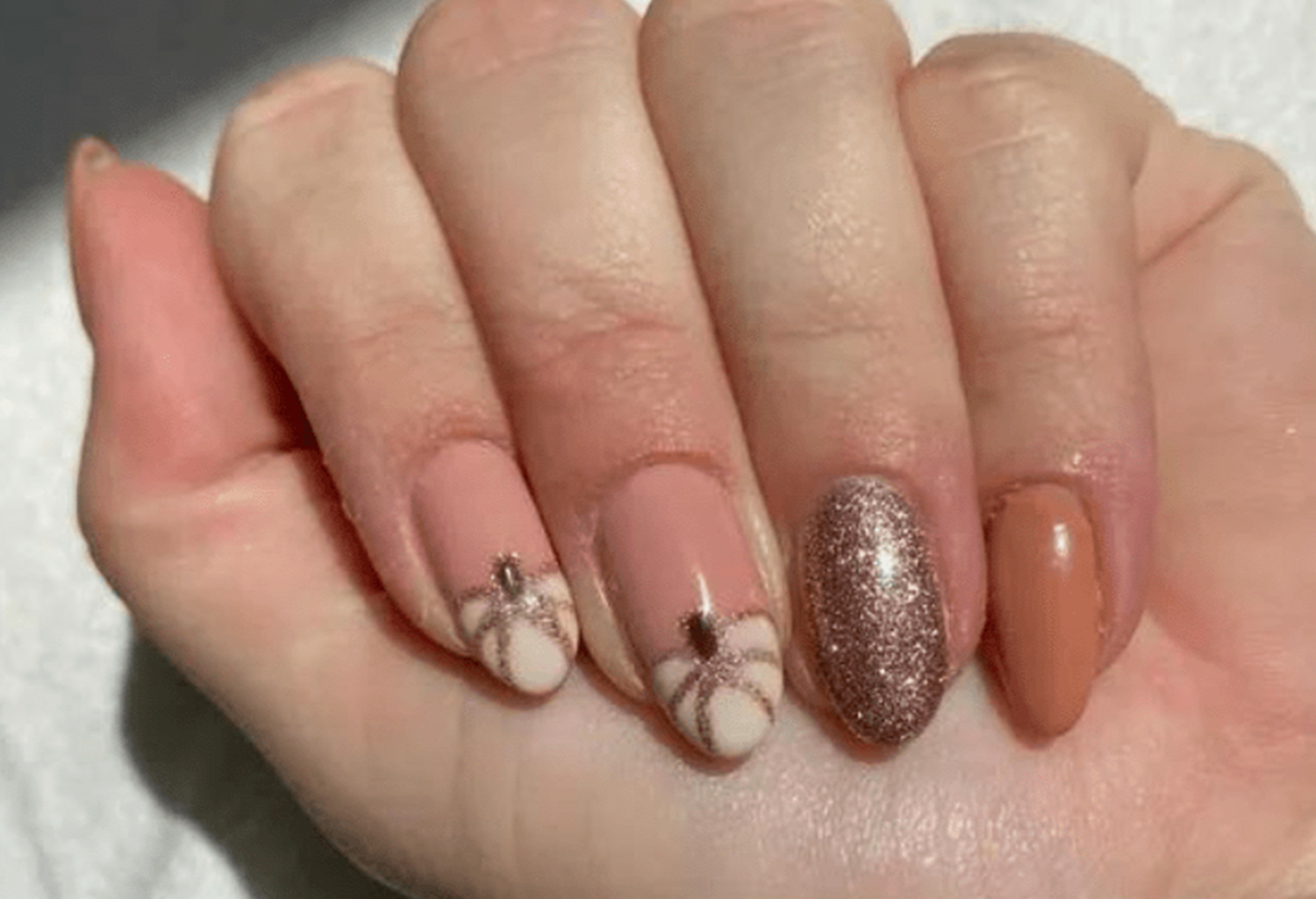 Hand with stylized nails in various designs; Pink with glitter accents, pumpkin motif and shiny copper-colored detail.