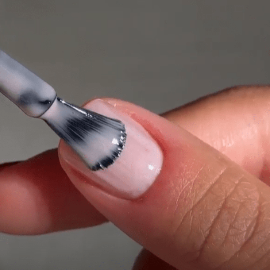 A close-up of a hand applying nail polish with a brush to a light pink nail.