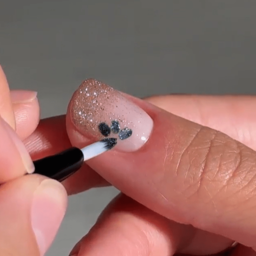 Brush paints black paw prints on a nail with shimmering, goldish nail polish.