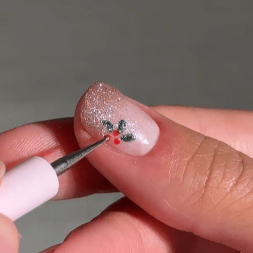 Close-up of a hand decorating a thumb nail with a Christmas design of holly leaves and red berries against a glittering background.