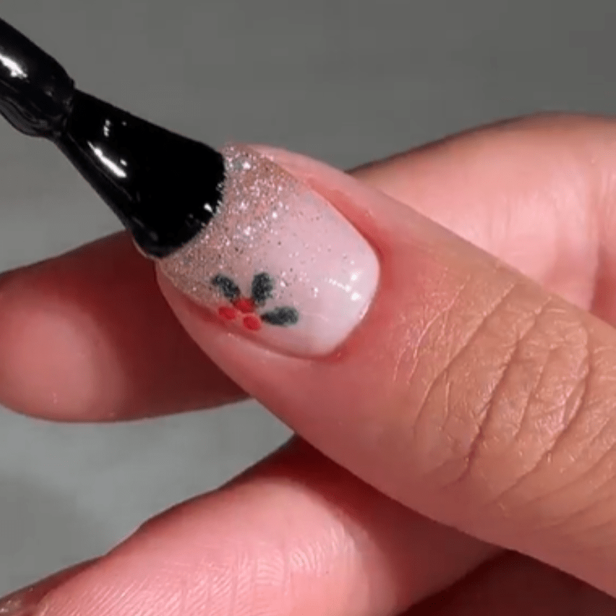 Close-up of a hand holding a nail with a shimmering silver lacquer and a design of green leaves and red dots.