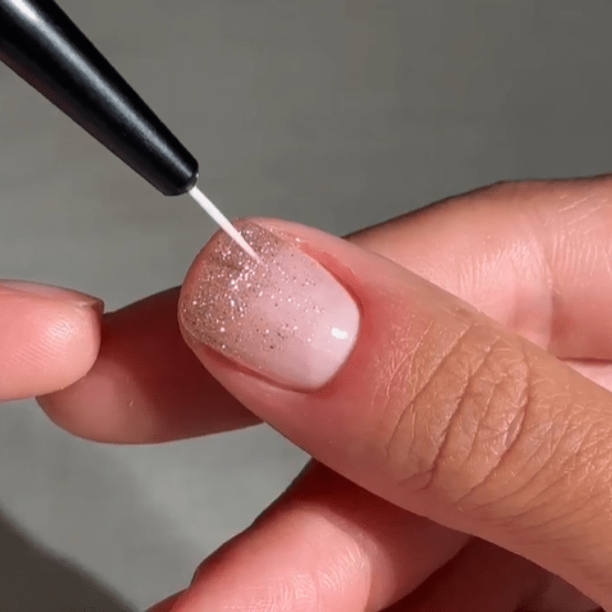 A hand showing a nail with pink nail polish and glitter, while a nail art brush applies a fine white line.