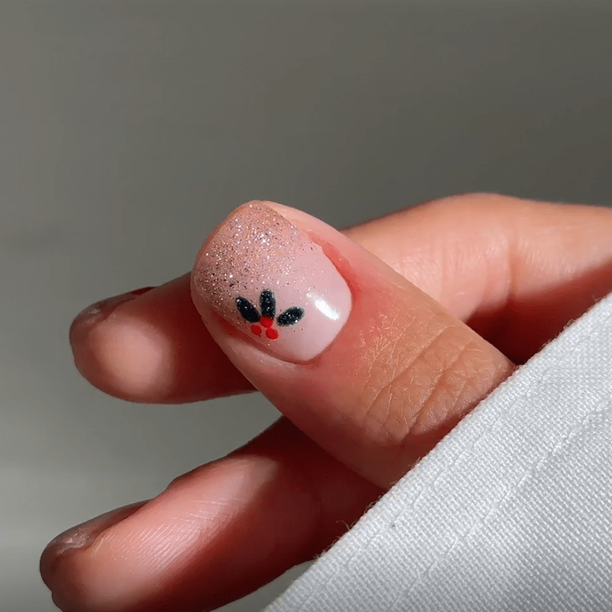 A thumb with a nail design of shimmering nude polish and a small floral pattern with black and red accents.