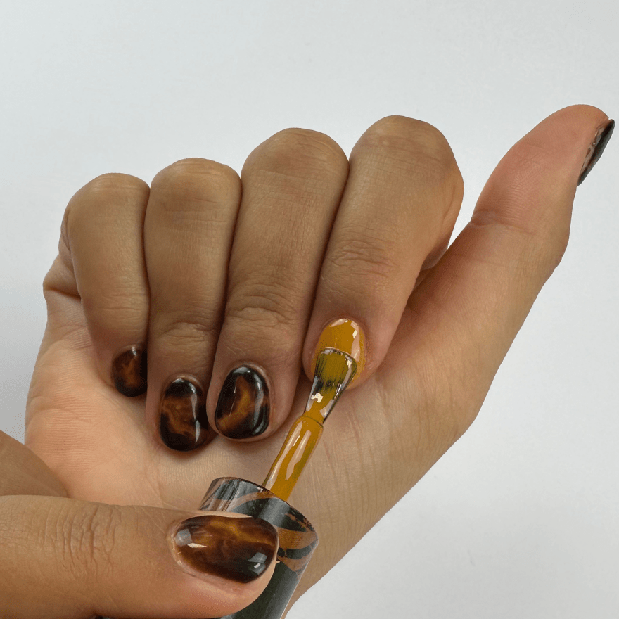 A hand with brown, marbled nail art, where the index finger is painted with a yellow nail polish brush.