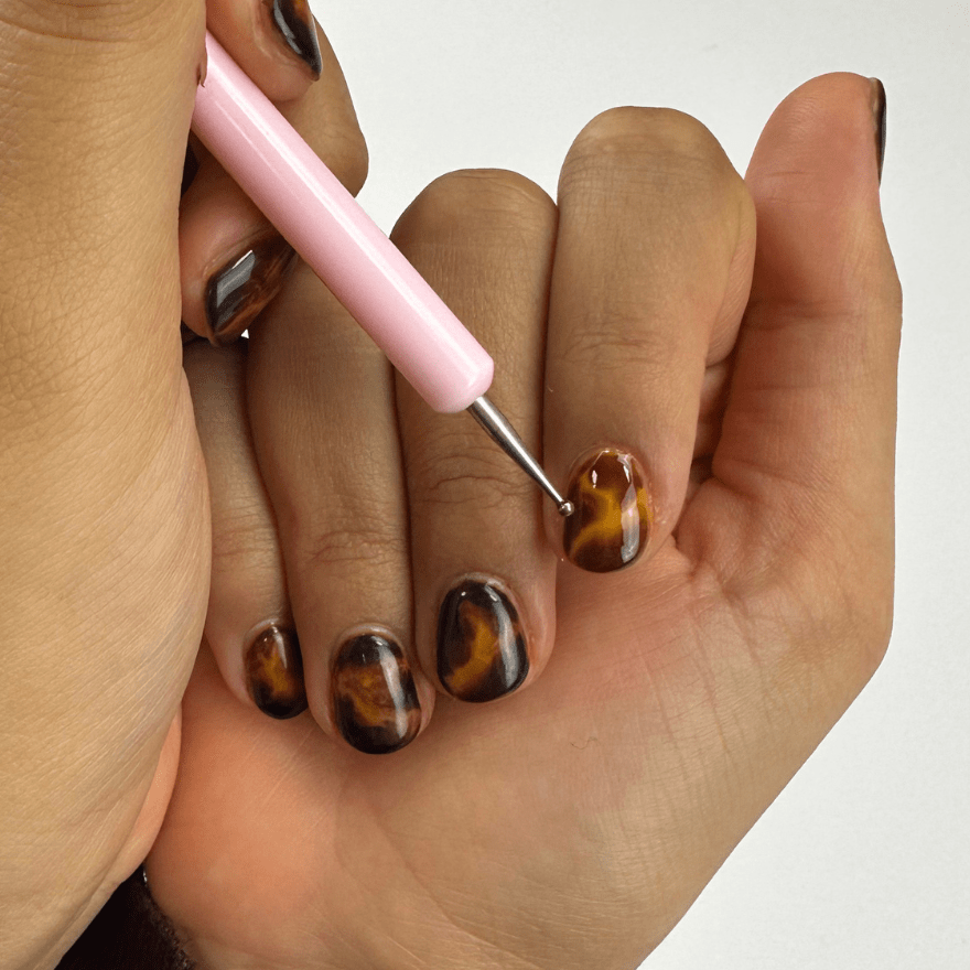 Close-up of hands with brown and black nail polish, with one of the nails being decorated with a nail polish tool.