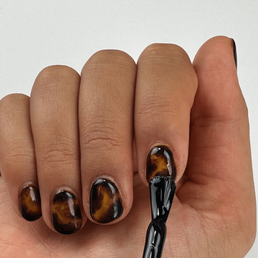 Close-up of a hand applying brown nail polish with a tortoise shell design to a nail with a brush.
