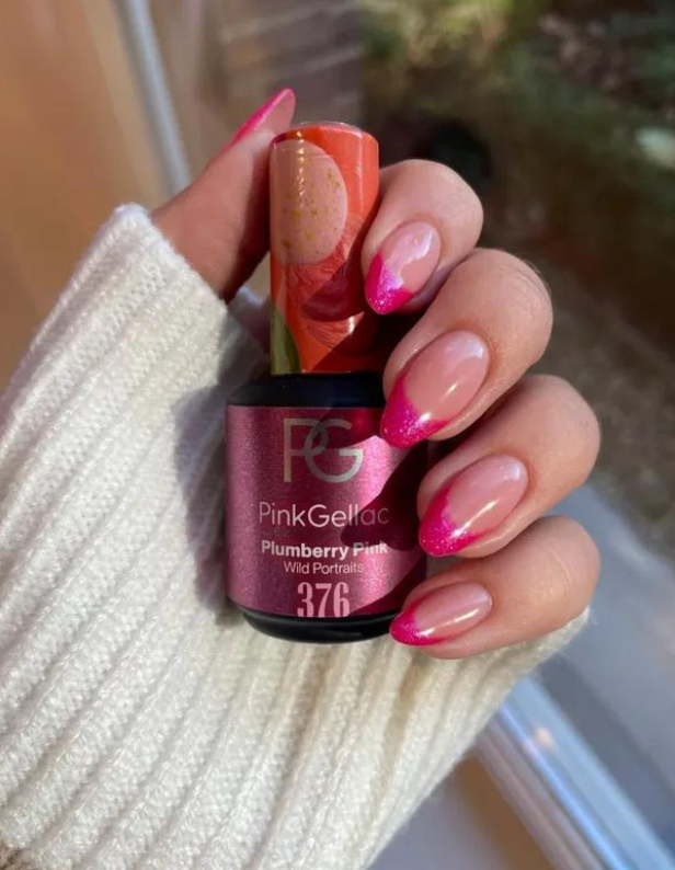 A hand in a white sweater holds a bottle of Pink Gellac nail polish with the color "Plumberry Pink", nails have a pink-french manicure.