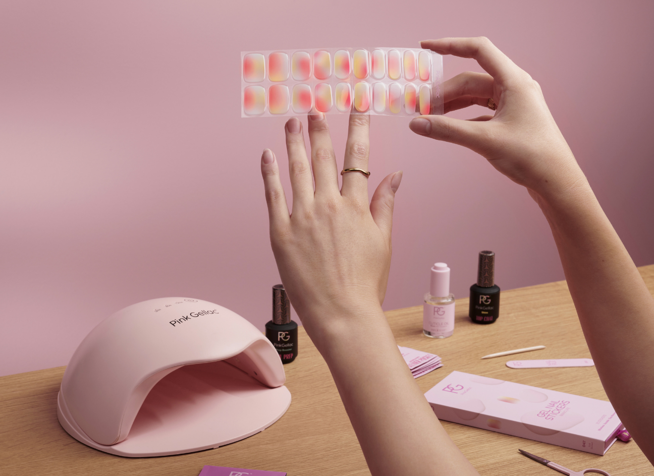 Hands holding a sheet of colored fake nails over a wooden table with nail products and a pink UV lamp.