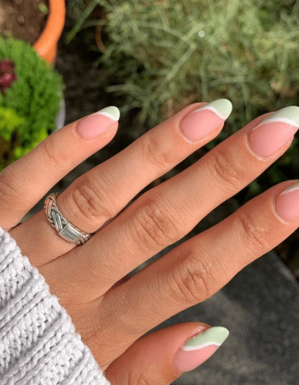 Close-up of a hand against a natural background, with a silver ring on the finger and nails with a pastel green and white wavy manicure.