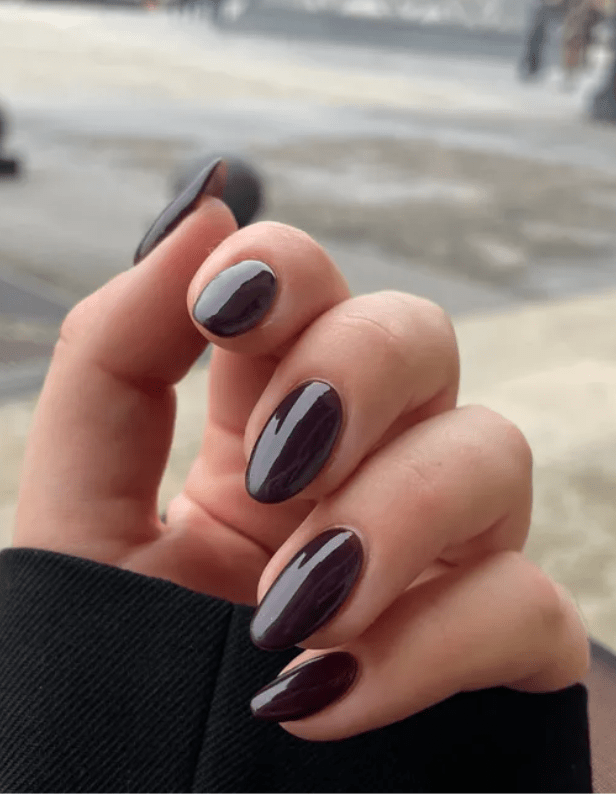 Close-up of a hand holding shiny, dark purple nail polish against a blurred background.