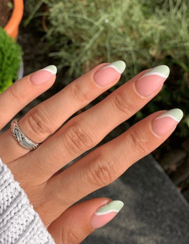 A hand with a silver ring and nails with pink lacquer and green, wavy tips against a background of green leaf.