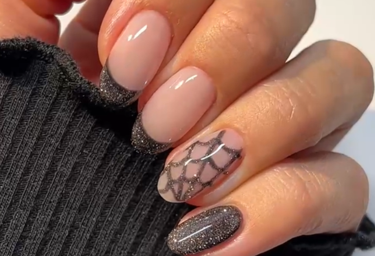 Hand with black glitter nail design, including a spider web pattern and black glitter tips, against black clothes.