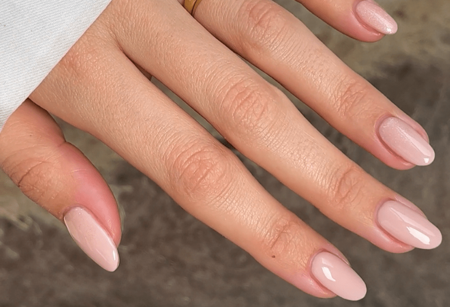 Close-up of a hand with pink nail polish and a gold ring on the index finger, on a gray background.