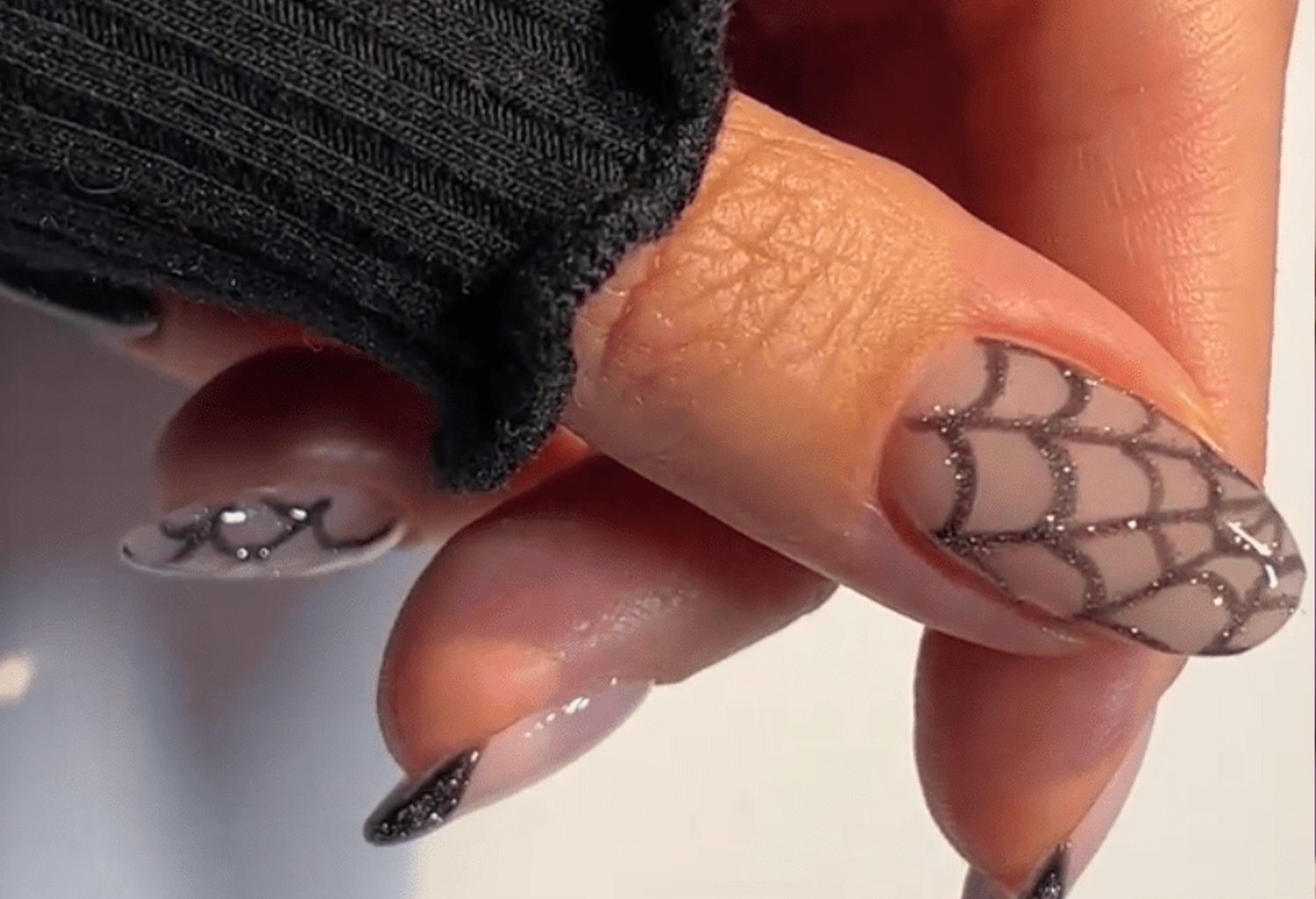 Nails with spider web design and glitter accents, against a dark sweater in close-up.