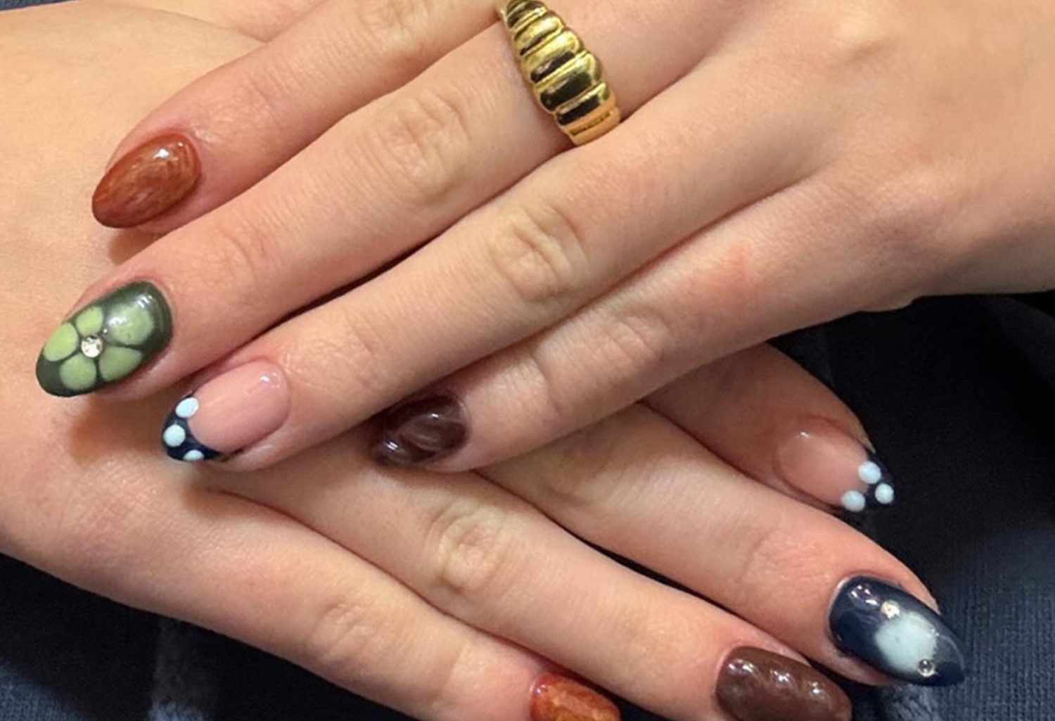 Two hands with all kinds of colorful and decorated nails, including floral and dot patterns, and a gold ring.