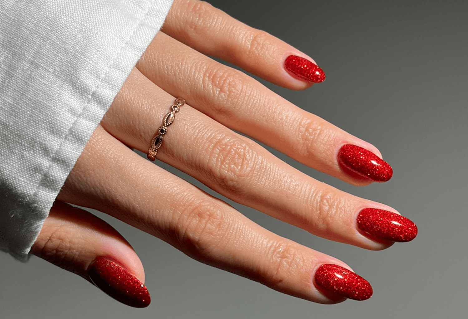 Hand with red glitter nail polish and a graceful gold ring on the finger, against a gray background.
