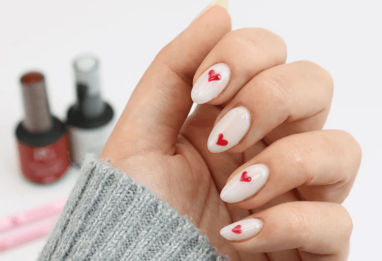 Hand with white nails decorated with small red hearts, next to some nail polish bottles.