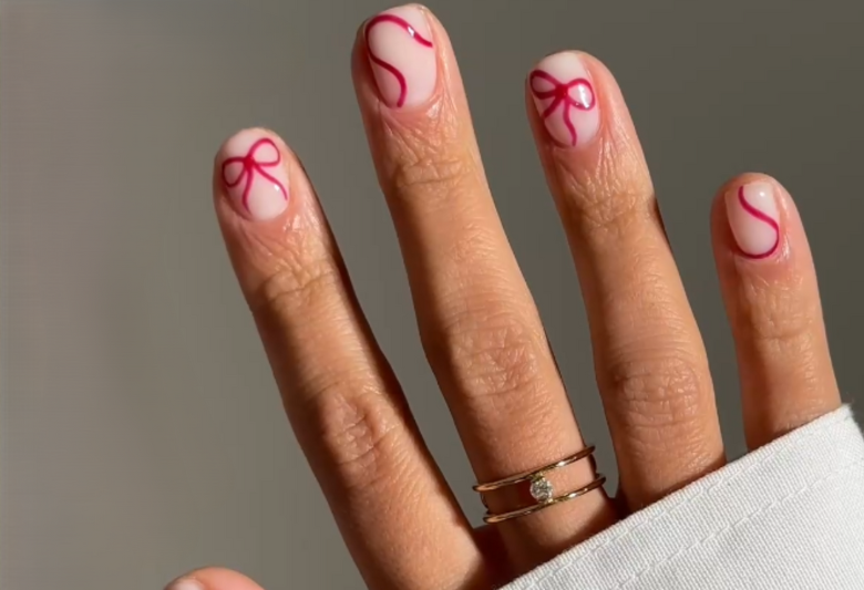 Close-up of a hand with painted nails with transparent base and pink bows as decoration, with a gold ring with a diamond on the ring finger.
